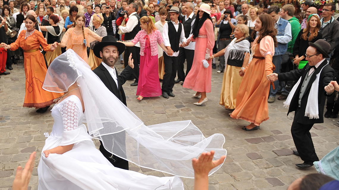 A Jewish couple is dancing in the middle of a circle of people holding hands. They are all outdoors.