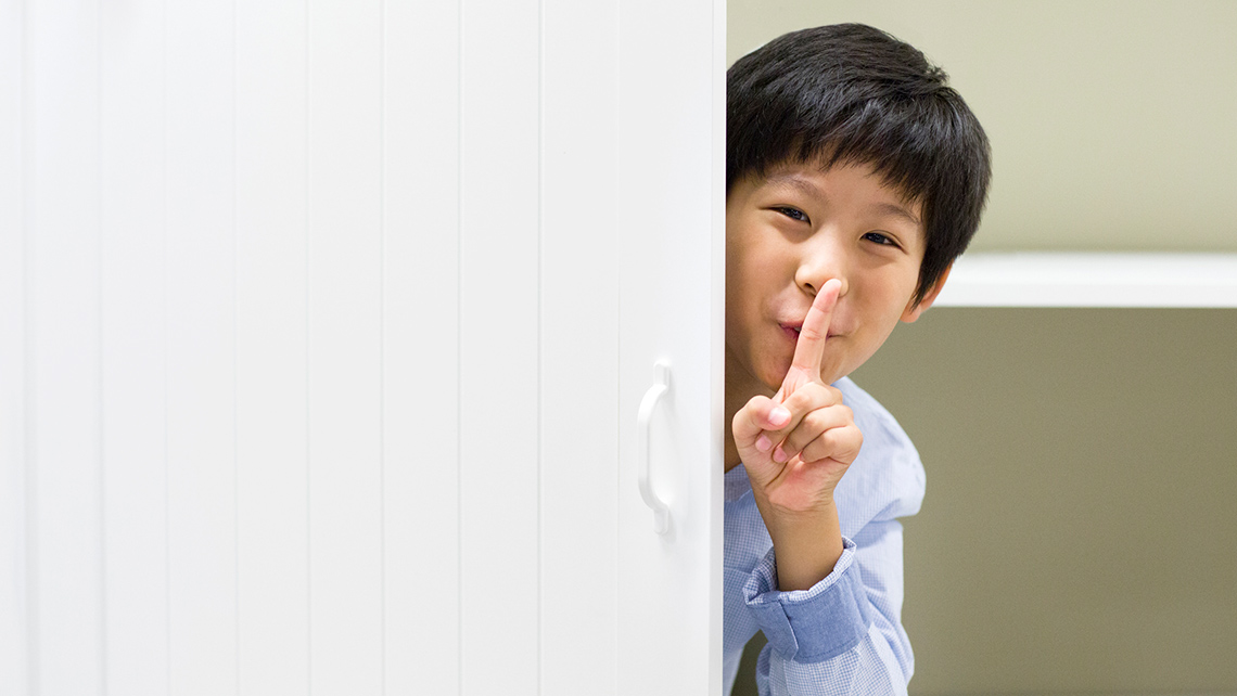 A child is leaning out from behind a door. Their finger is raised to their lips and they are signaling to be quiet.