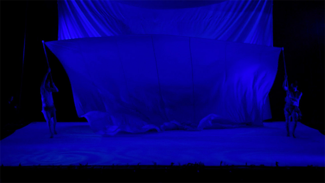 2 dancers are on opposite ends of the stage, each holding a stick. The sticks are attached to a large piece of fabric.
