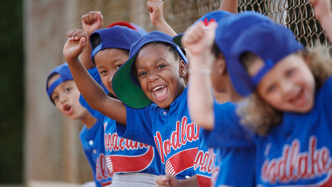 A group of children, in their uniforms and sitting on a bench, are cheering at a baseball game.