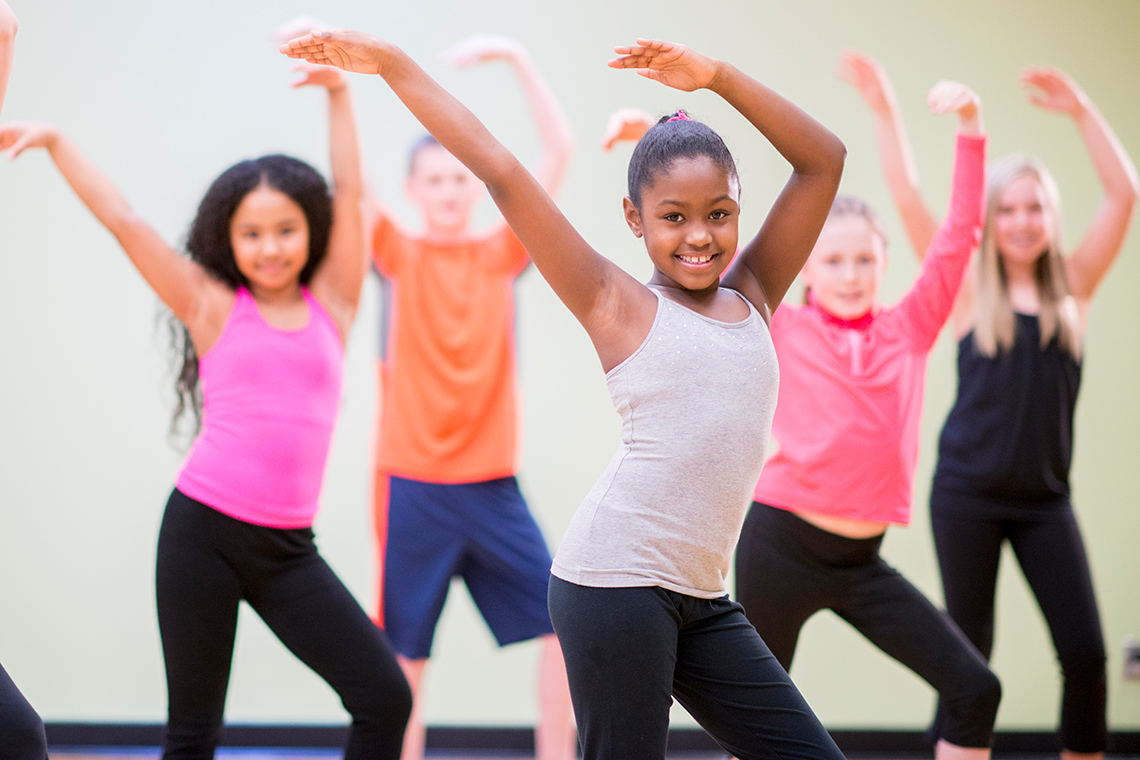 A group of 5 young people wave their arms above their heads as they dance indoors. One person is dancing in front of the other 4 people.