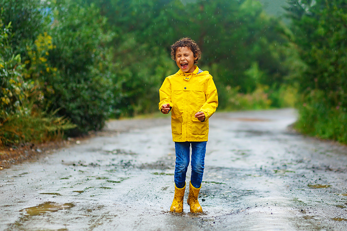 A young person, wearing a bright raincoat and rainboots, laughs and dances in the rain on a muddy dirt path that is surrounded by trees.