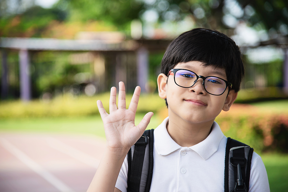 A young person, wearing a backpack and glasses, holds their hand up in a wave gesture as a form of greeting outdoors on a sunny day.