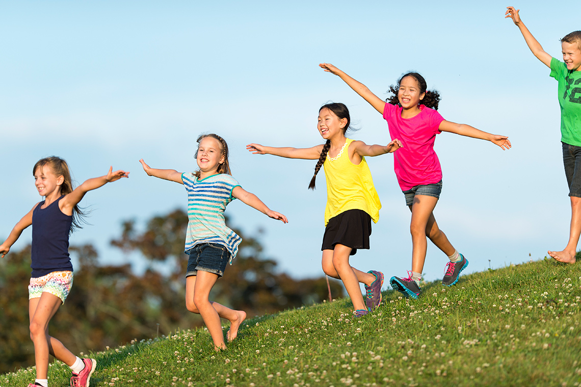 Five young people, with both their arms raised level with their shoulders, walk or dance down a grassy hill outdoors on a sunny day.