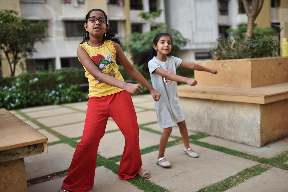 Two young people, standing beside each other, swing their hips and move both their arms to the right as they dance outside on a patio.
