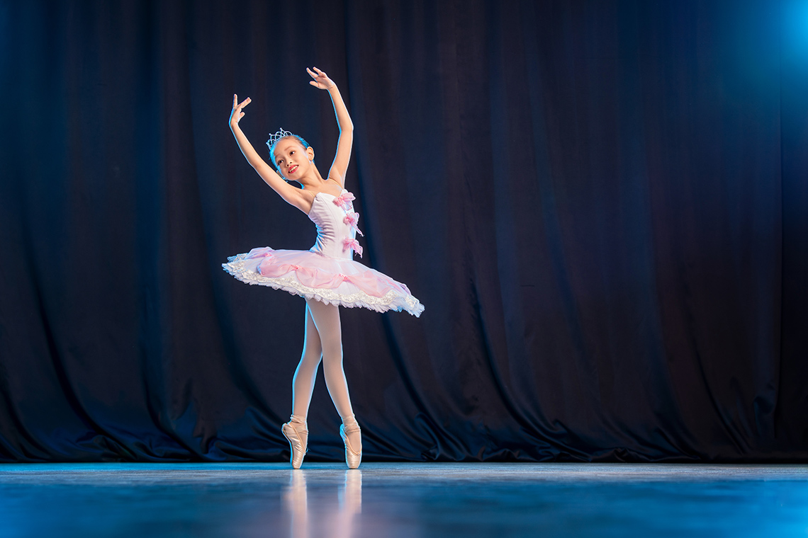On a stage, a young ballerina, looking to their right, stands on their toes with their arms raised in a curve above their head.