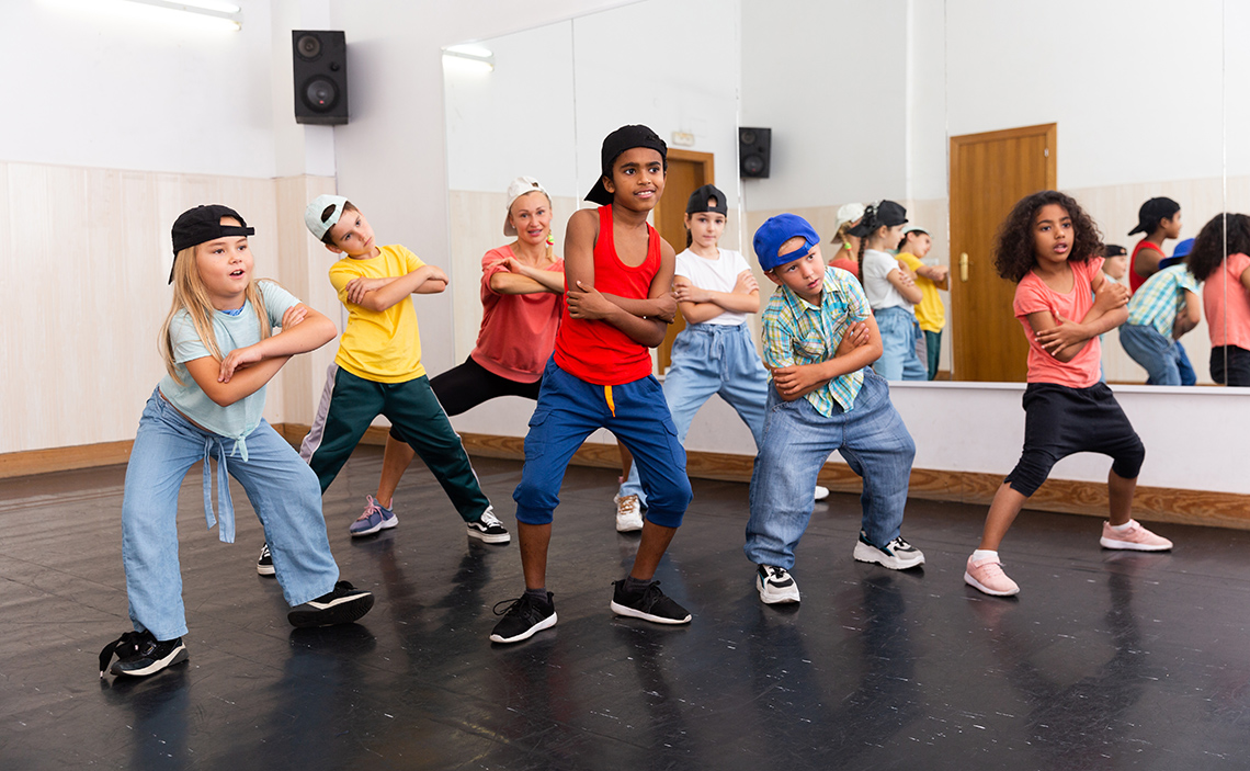 Seven students stand with their legs apart, bent forward at the waist, and arms crossed in front of them, practice dance moves in a studio.