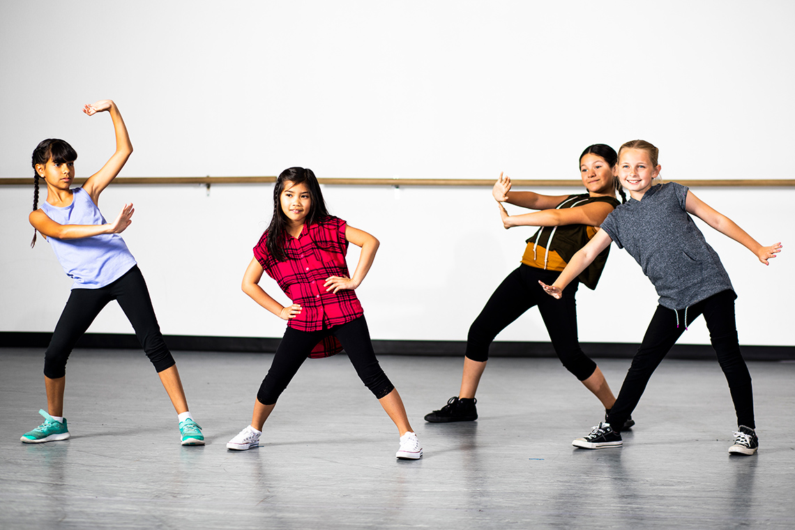 Four students practice hip hop dance moves indoors in a dance studio with arms above their head, stretched out to the side or hands on hips.