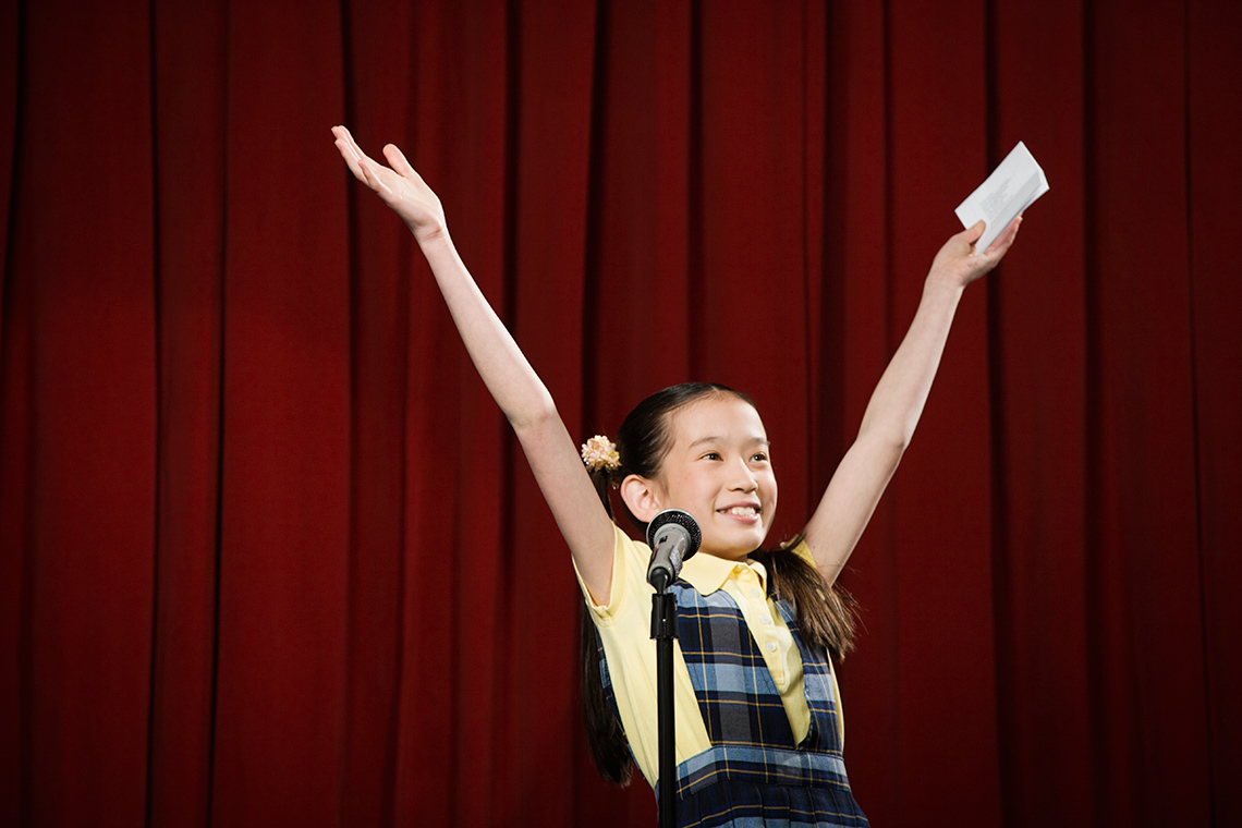 A young person stands on a stage behind a microphone, hands raised in the air triumphantly above their head, holding a piece of paper.
