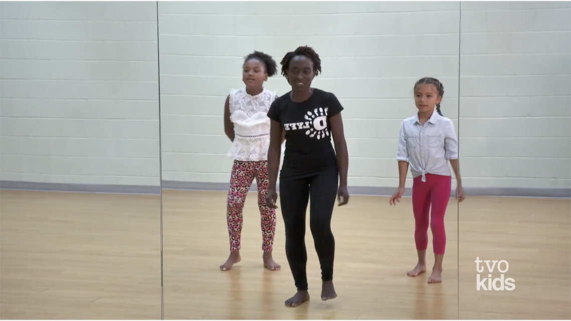 An instructor and 2 students shuffle their feet with their knees slightly bent in front of a wall-length mirror indoors in a dance studio.