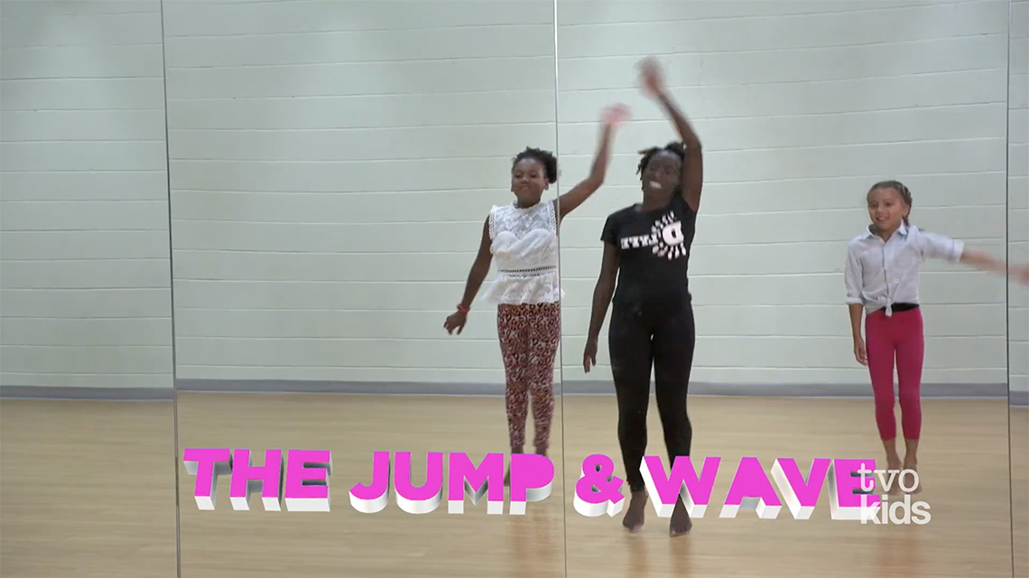 Three young dancers jump up and down and wave their arms. They are dancing in front of a wall-length mirror indoors in a dance studio.