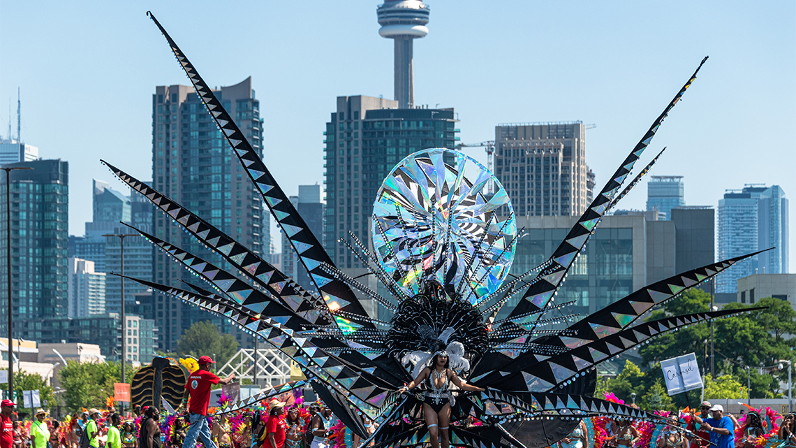 A Toronto Carnival dancer moves down the street wearing a float that resembles a bird with big wings. It has a big headdress and wheels.