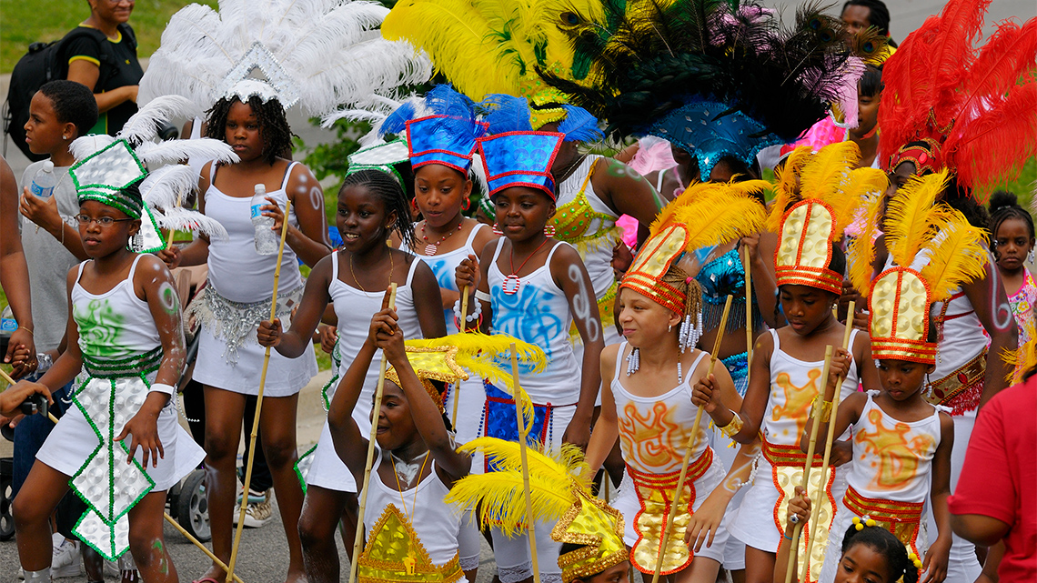 Young people at the Toronto Carnival parade move down the street wearing feathered headdresses, sequined outfits, and holding long sticks.
