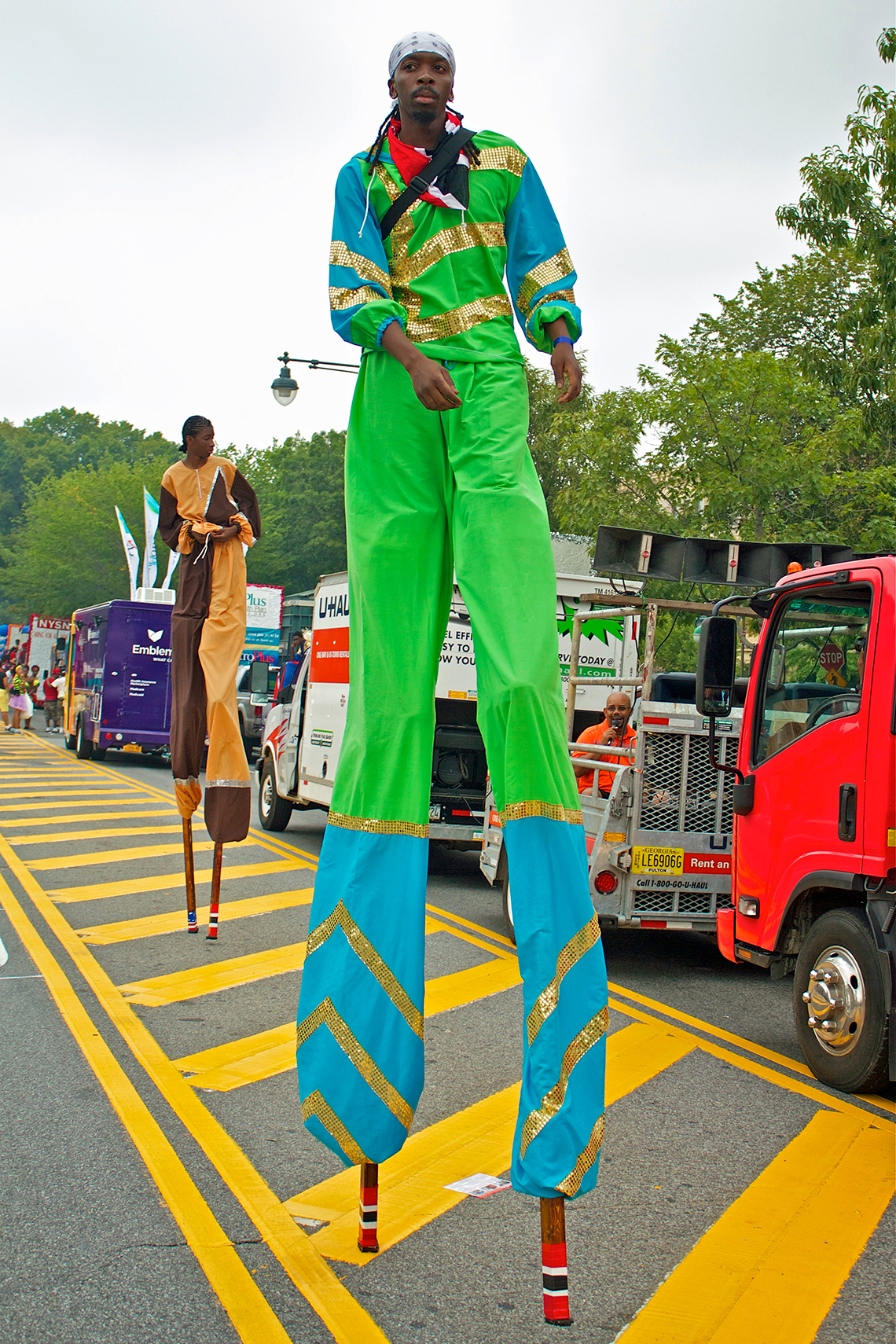 Two Toronto Carnival dancers move along the street on long, tall stilts, wearing bright coloured pants, tops, and hats.