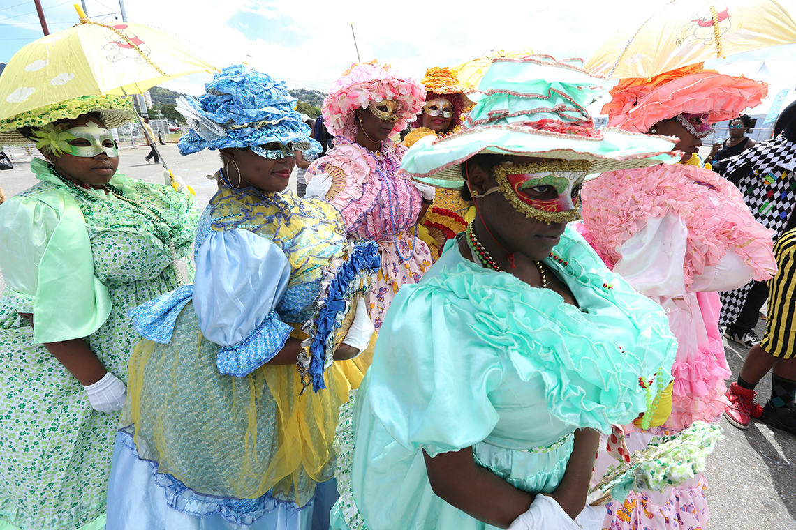 A group of Carnival masqueraders dressed in colourful ball gowns wearing big colourful hats, carrying parasols and fans, and wearing masks.