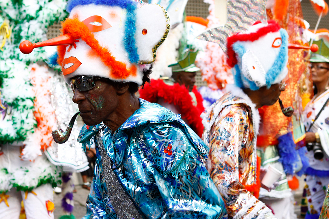 A Carnival dancer wears sea-coloured pants and jacket, a hat with a face and fish on top, carries a cane, and holds a pipe in their mouth.