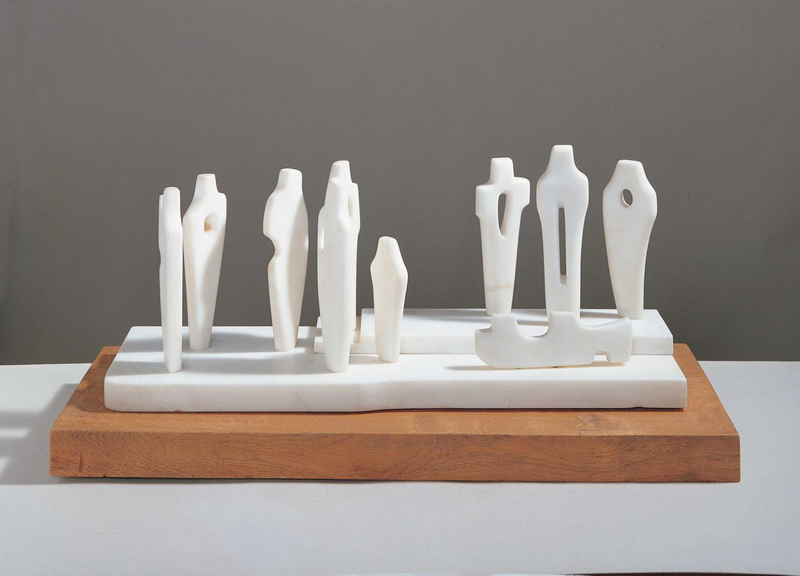 A white sculpture of 8 human-like figures, 7 of which are tall, 1 is short. 3 are on a raised section. A non-human figure is lying down.