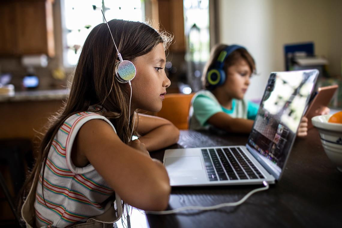 Two students sit at a table indoors in front of their own laptop, each of them wearing headphones that are plugged into the laptops.