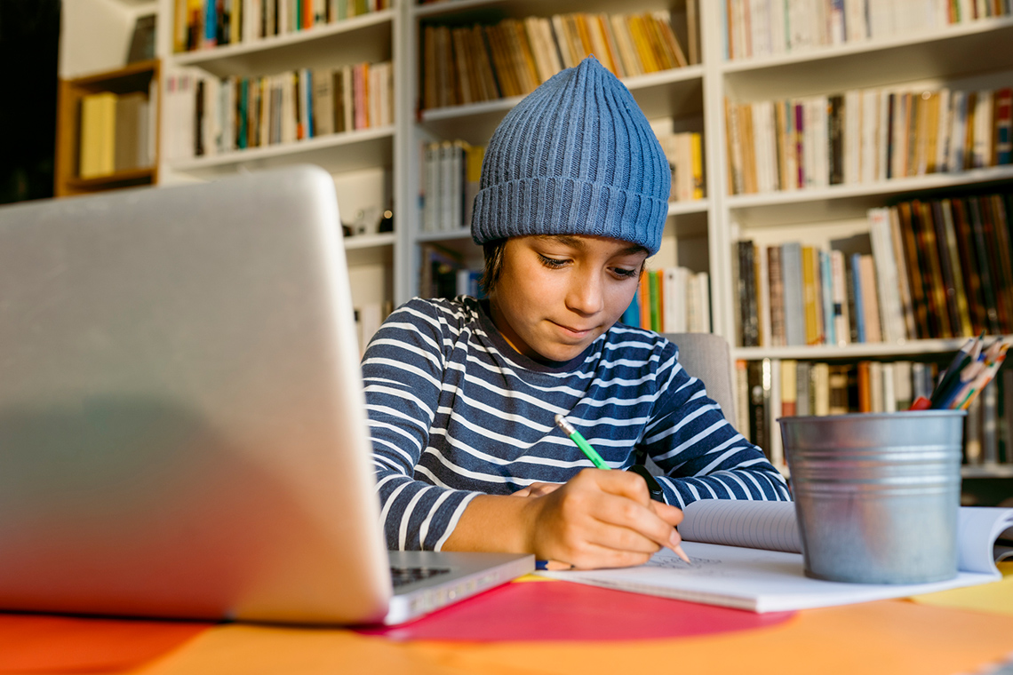 A student sits at a table in front of a wall of bookshelves, writing in a notebook with a pencil, with a laptop open in front of them.