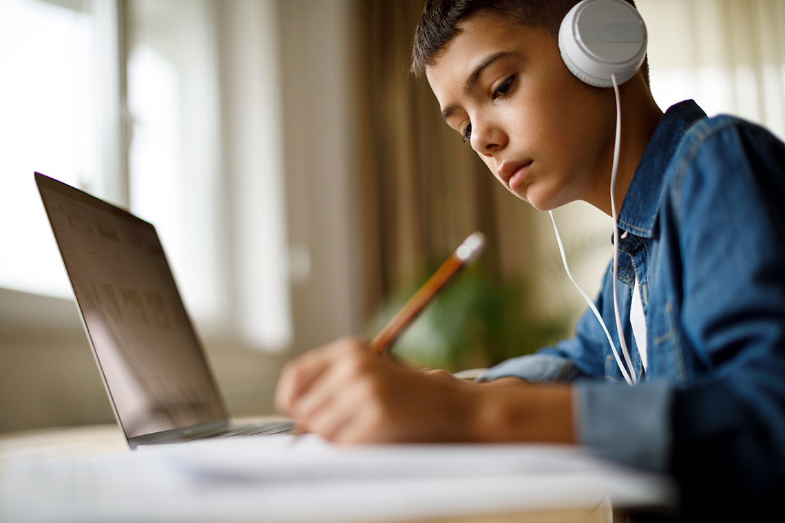 A student, wearing headphones and a long-sleeved shirt, sits at a desk indoors writing with a pencil in a notebook beside an open laptop.