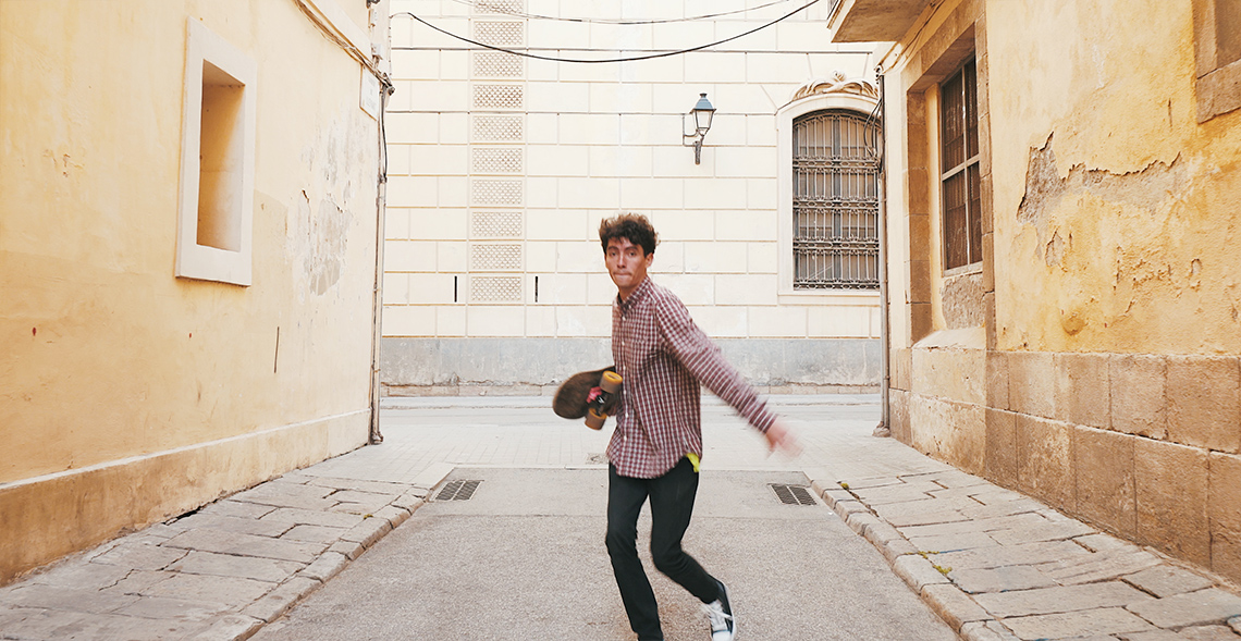 Dancer looking forward A dancer, their body turned to 1 side, dances on a street outdoors between 2 buildings, holding a skateboard in 1 arm, looks at the camera.