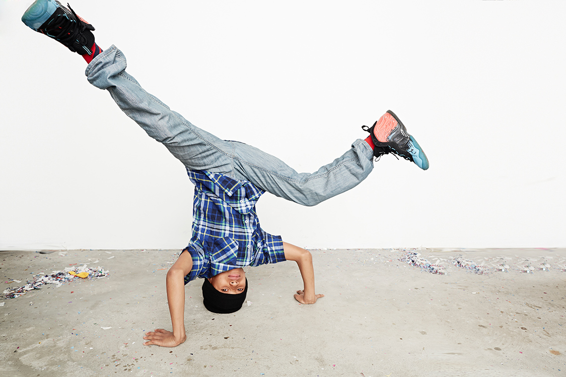 Breakdancing A young dancer performs a breaking dance move, a headstand with their hands on the ground for balance and legs apart outdoors on concrete.