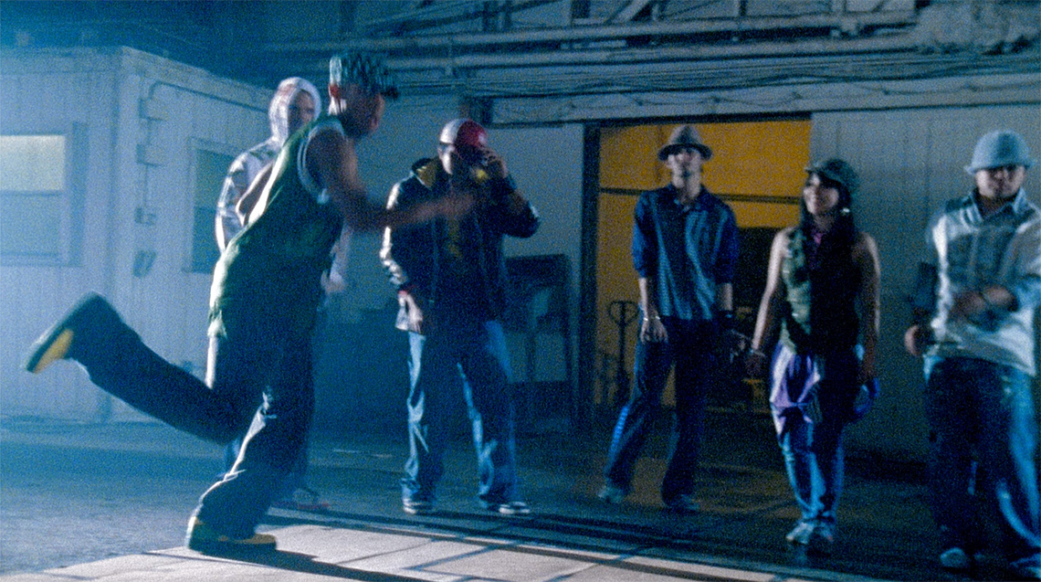 A group of six dancers, all wearing comfortable and loose clothing such as t-shirts, sweaters and pants, dance indoors in a warehouse.