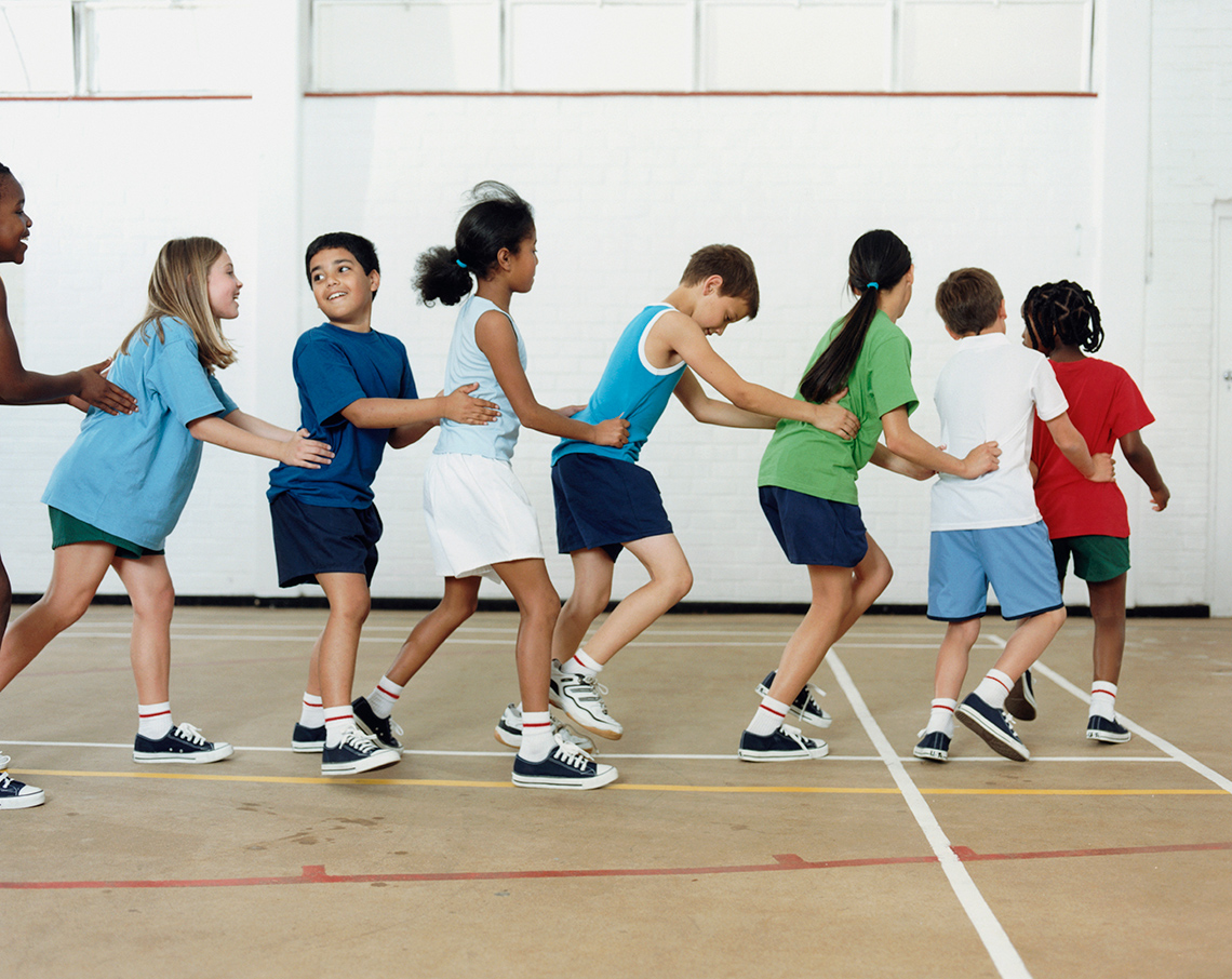 Conga line A group of eight young people, all wearing t-shirts, shorts and running shoes, move forward as they dance in a conga line indoors in a gym.