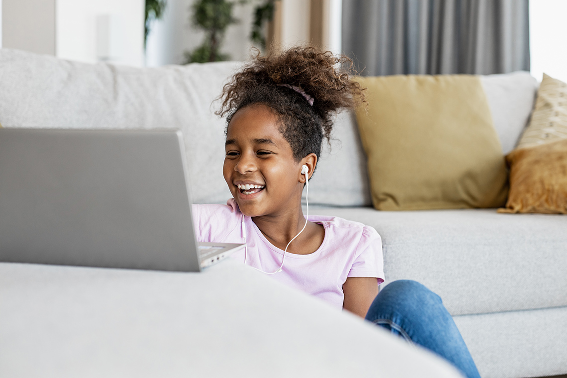 Investigate further A student sits on the floor in front of a couch indoors, with a laptop open on top of the couch. They are wearing ear buds and are laughing.