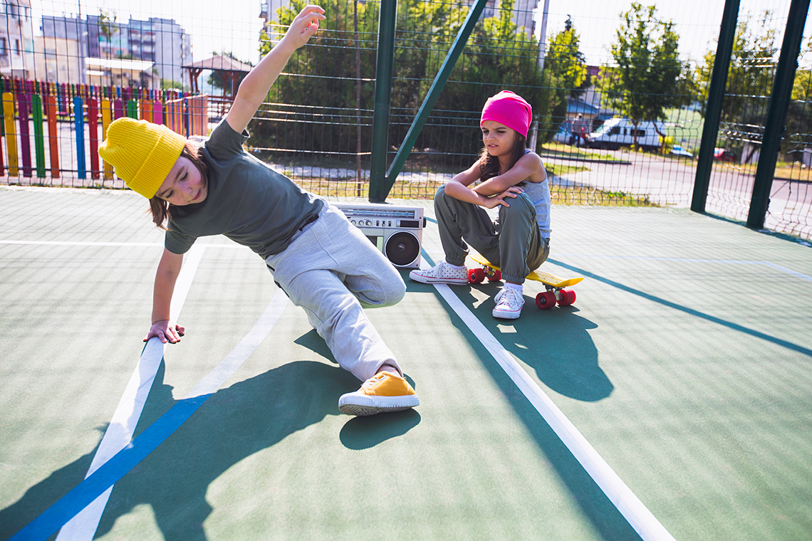 Two young dancers are on a sunny outdoor court. One dancer sits on a skateboard beside a large radio, 1 performs a breaking dance move.