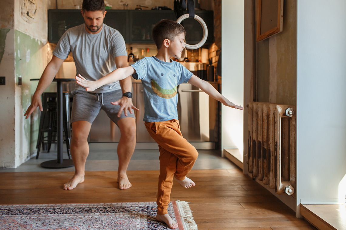 Dance video An adult and a student dance indoors in a hallway. The student stands on 1 leg, other leg crossed and arms out, the adult bends their knees.