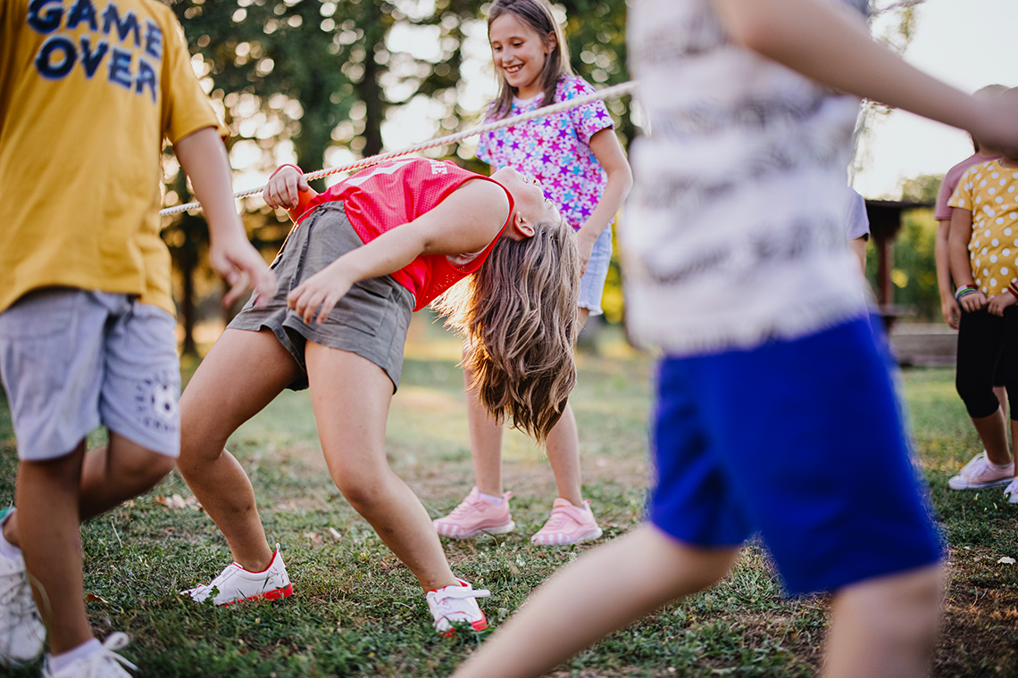 Two adults are holding a ribbon in a straight line off the ground. Two children bend backwards to walk under the ribbon.