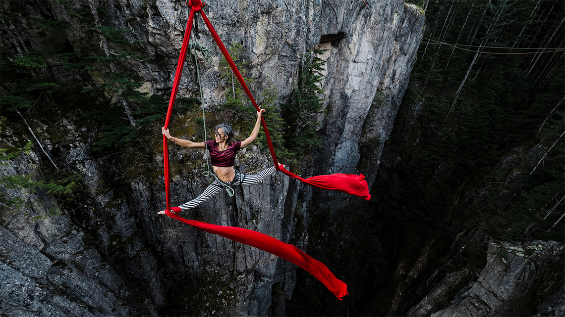 An acrobat adult dancer is suspended by an aerial silk from a tall rocky cliff.