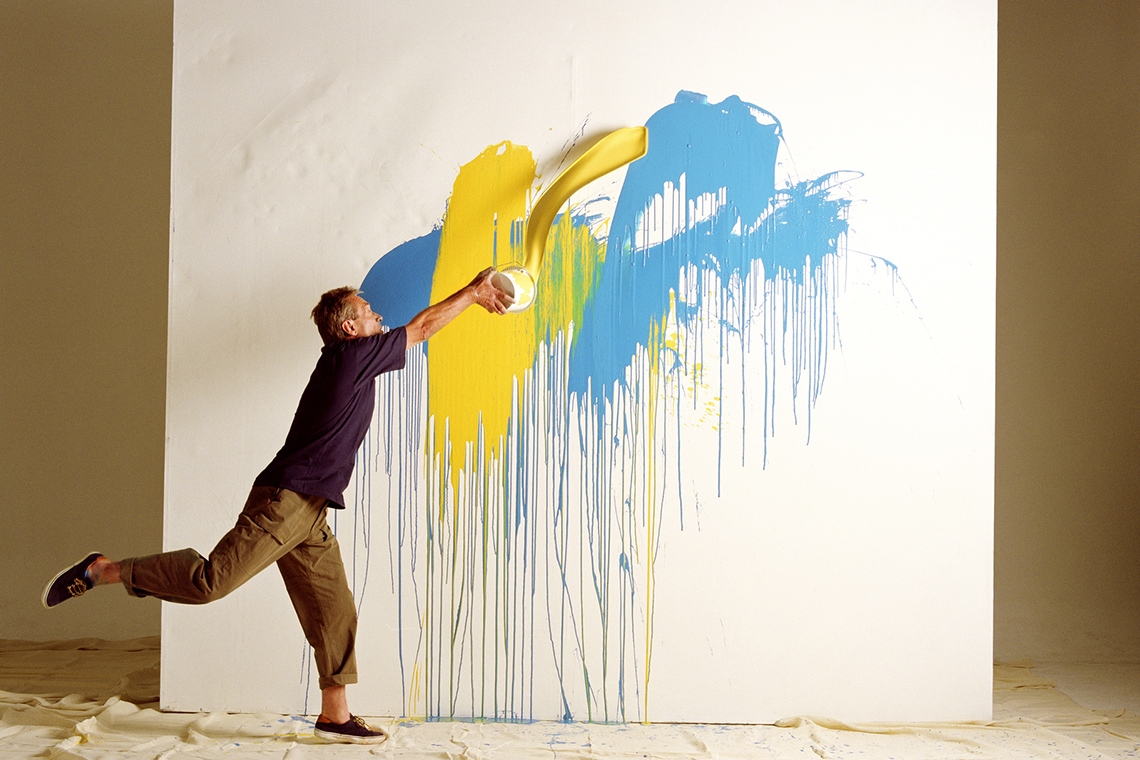 An artist is splashing paint on a large canvas that is put against a wall.