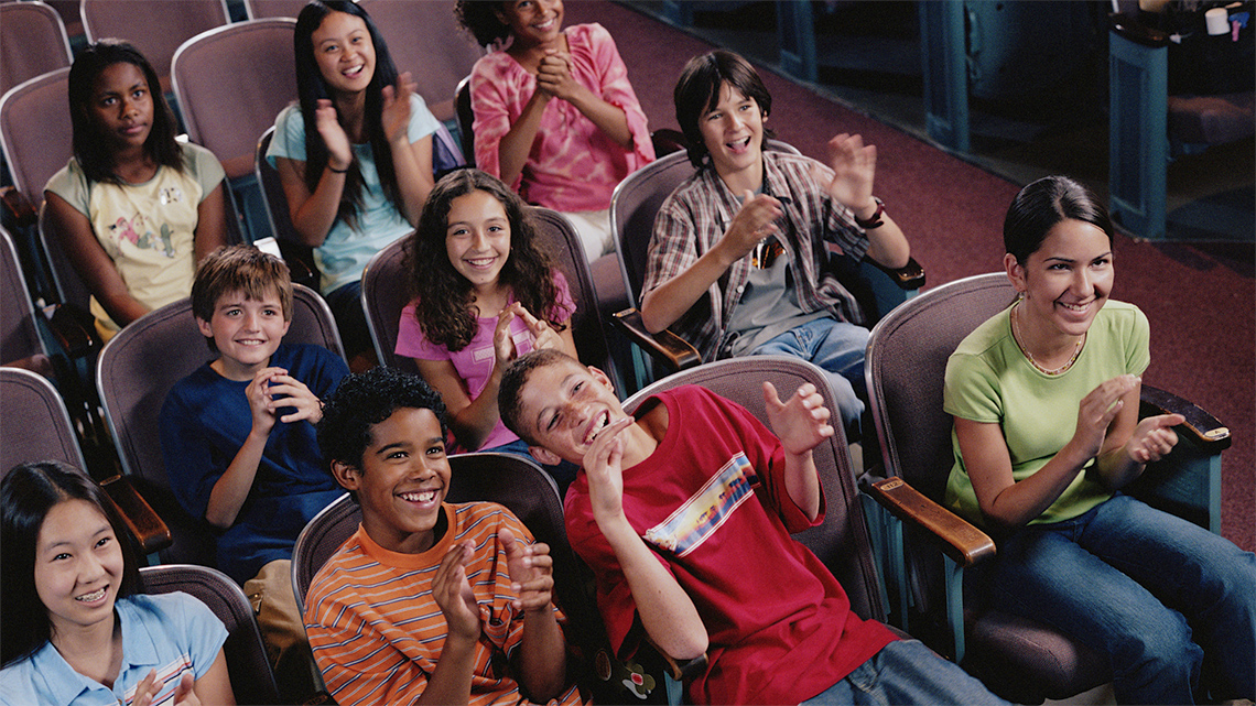 A group of students and an adult sitting in the theatre. They are all looking up at the stage.