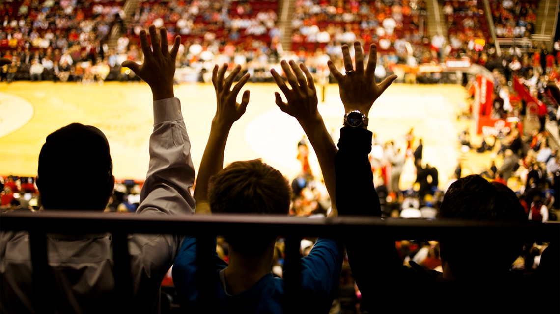 An image of a sporting arena showing the backs of several fans with their arms in the air.