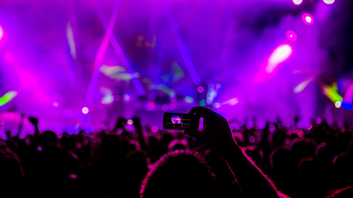 An image of a music concert showing the back of the audience. Many have their cameras raised. The room is dark and smokey.