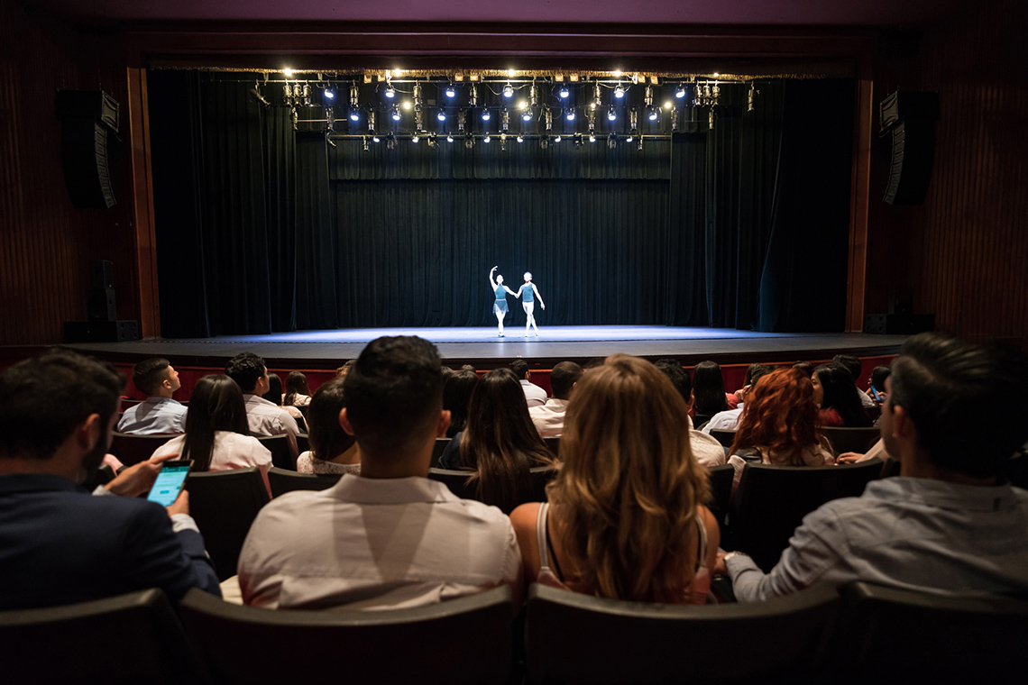 Two ballet dancers are on a stage in the background. In the foreground is the audience’s back.