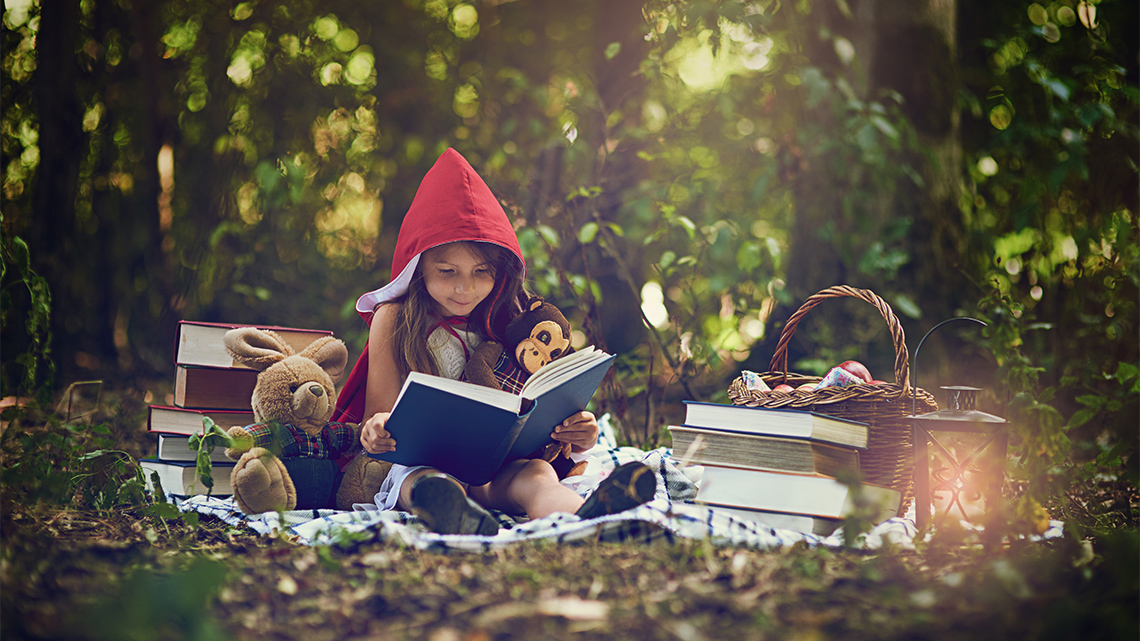 A child wearing a hooded cape. They are surrounded by books and reading a story out loud to stuffed animals.
