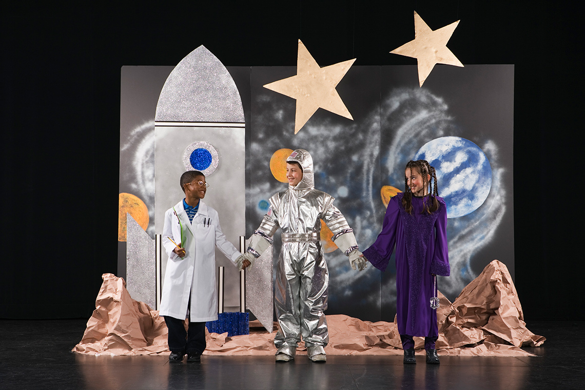 Drama performers standing in front of outer space themed scenery that includes stars, planets, and a rocket ship