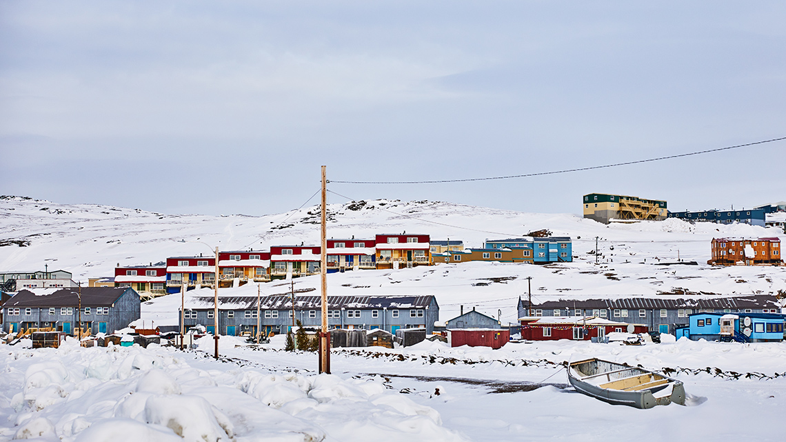 Houses surrounded by snow in Iqaluit, Nunavut. There are mountains in the background. A boat is parked in front of the houses.