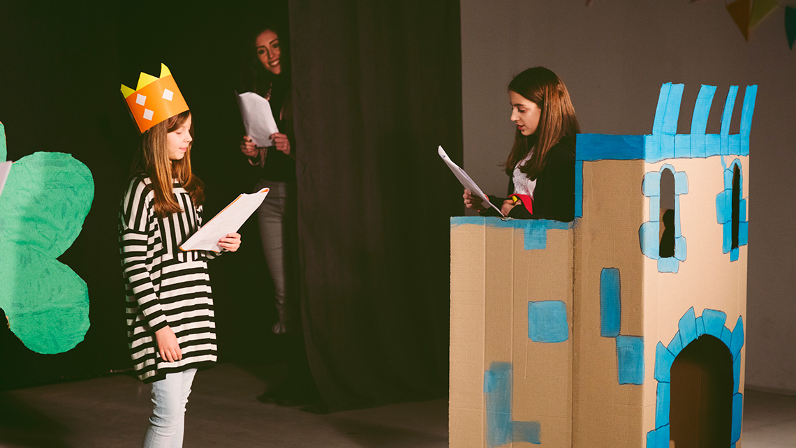 Two children wearing crowns rehearsing on stage with scripts in their hands.
