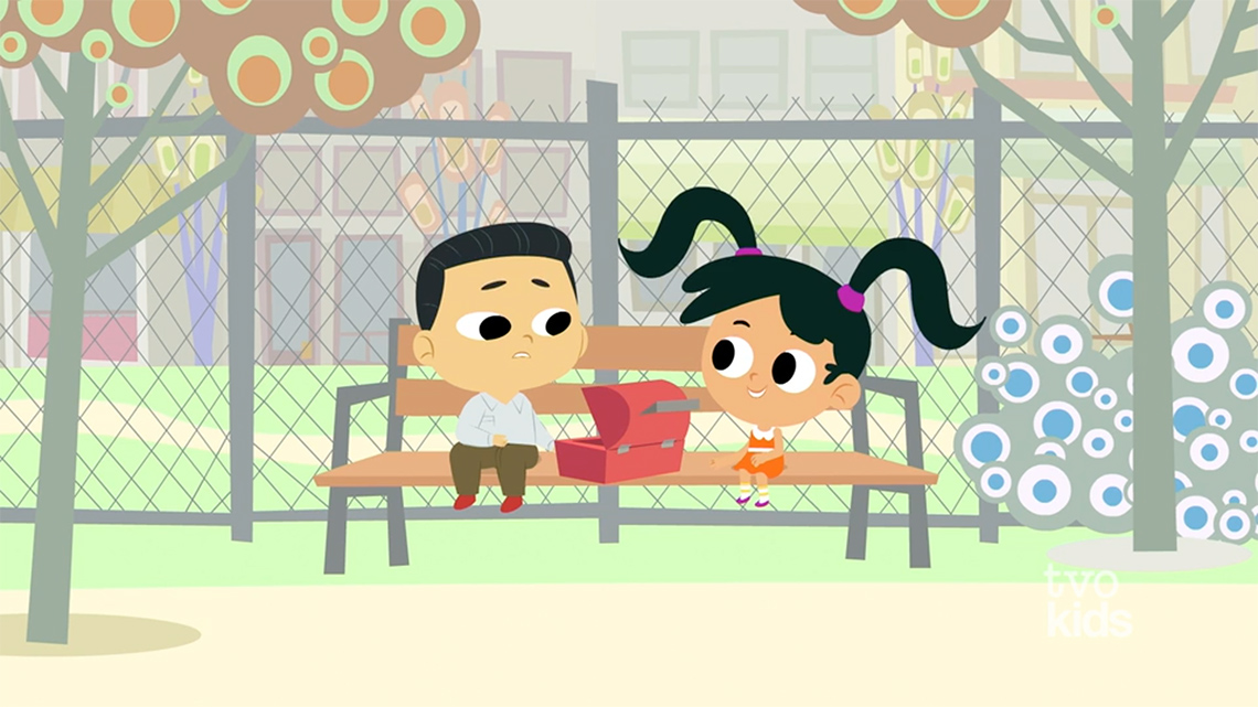 A screenshot from the video 16 Hudson: Picture Perfect with two children sitting on a bench outside. There is a lunchbox between them.
