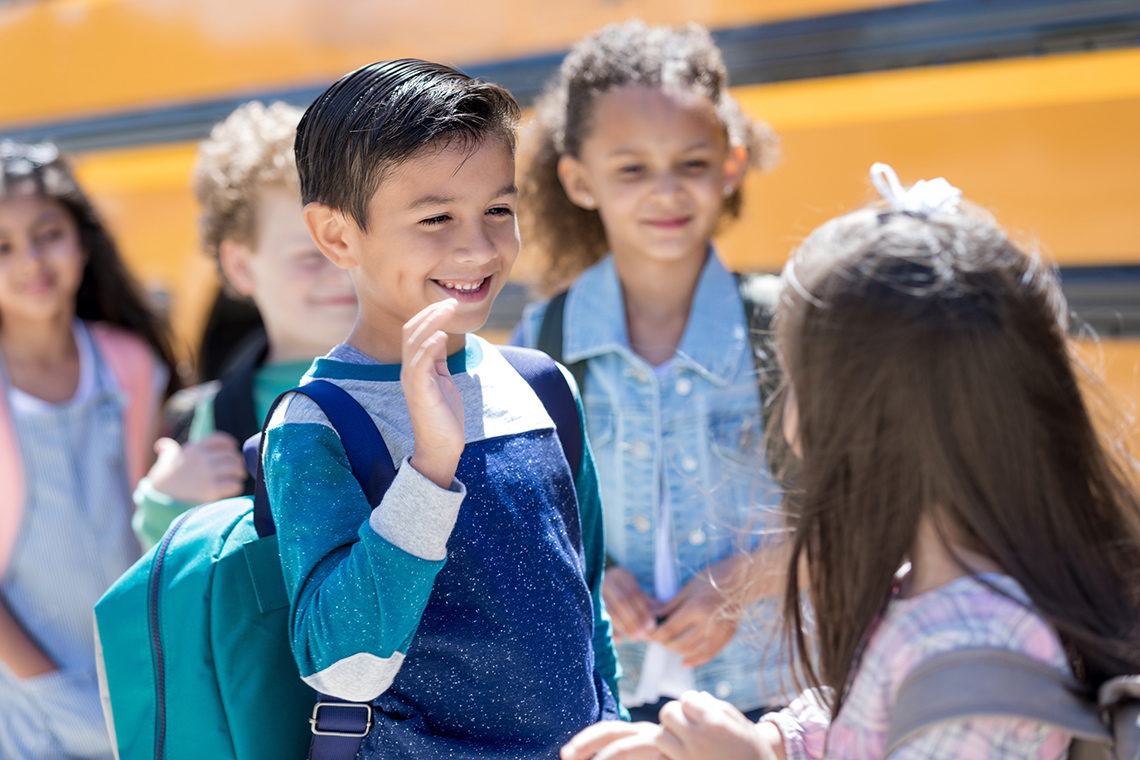 A child waving to another child near a school bus.