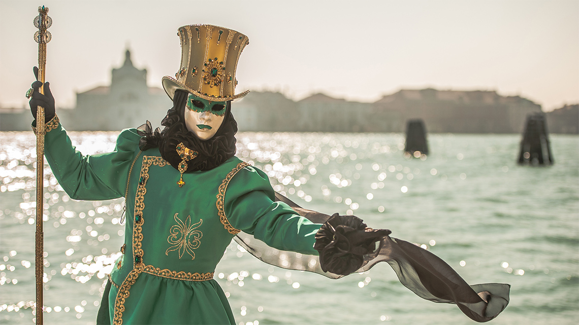 Person with a mask, a scepter, and a Venice carnival costume posing near a canal in Venice
