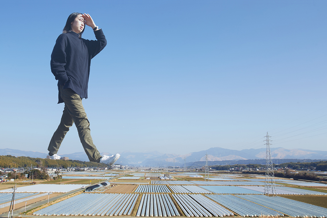 A giant person is walking outside through an open area of industrial buildings.