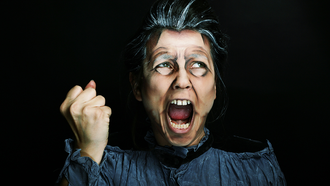 A person wears dark clothing and dramatic stage makeup as they shout and hold their fist up.