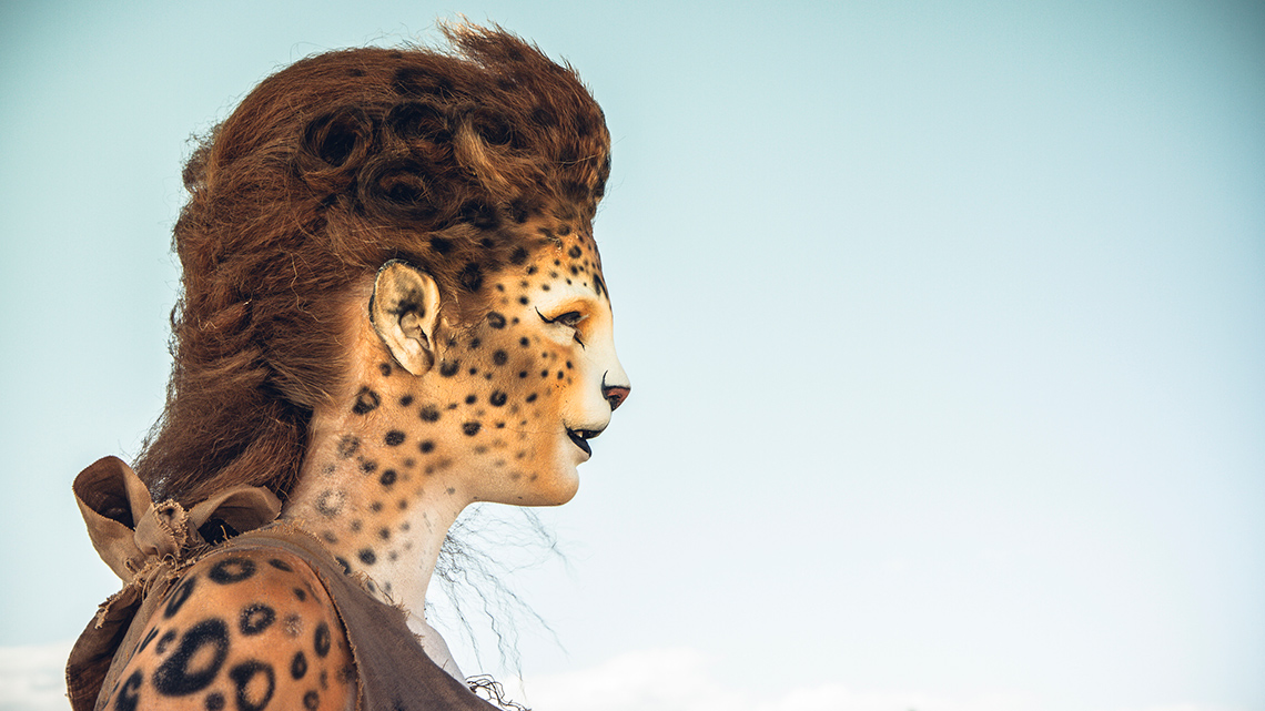 A person stands in a desert, wearing makeup on their face and arm to resemble a cheetah.