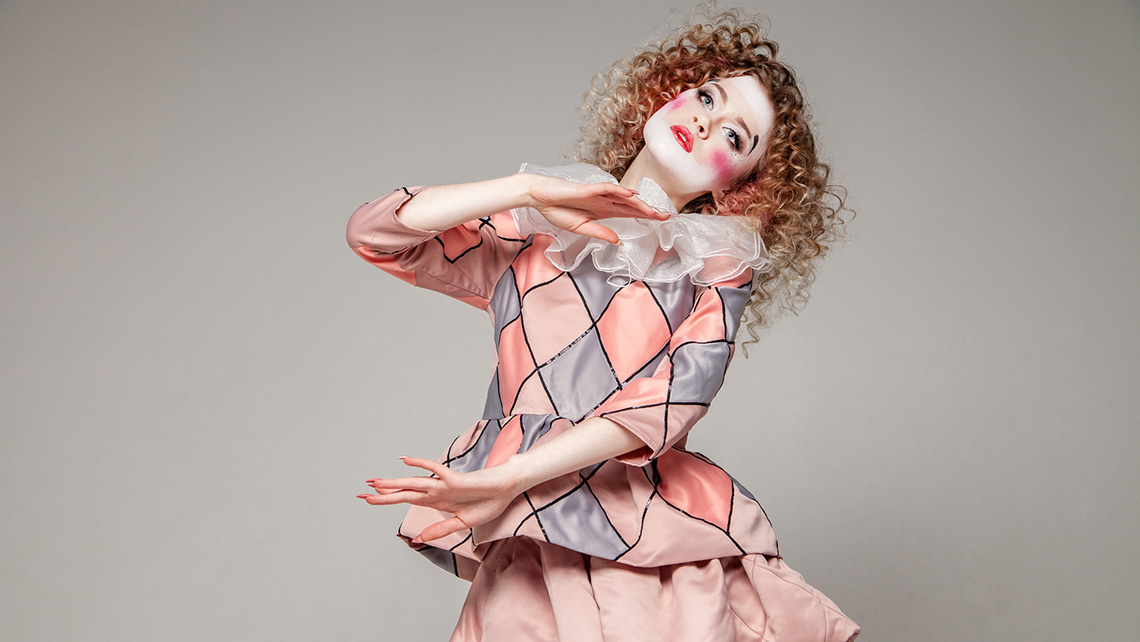 A court jester wears clown makeup and poses with two hands up and their head tilted.