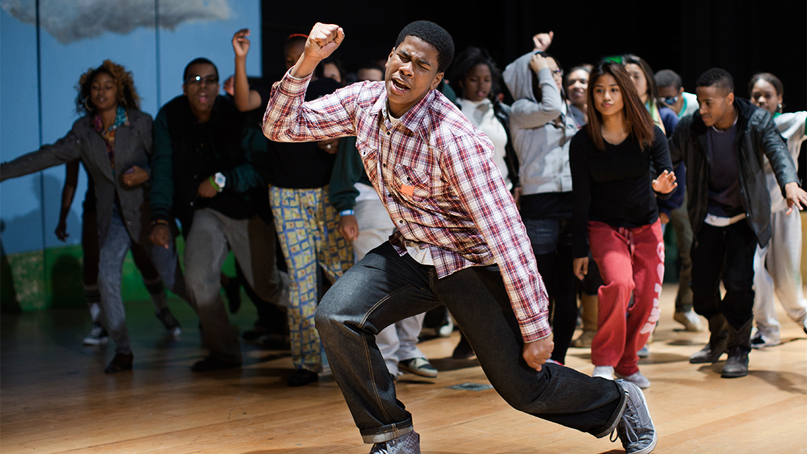 A person dancing on a stage. A crowd behind cheers them on. 